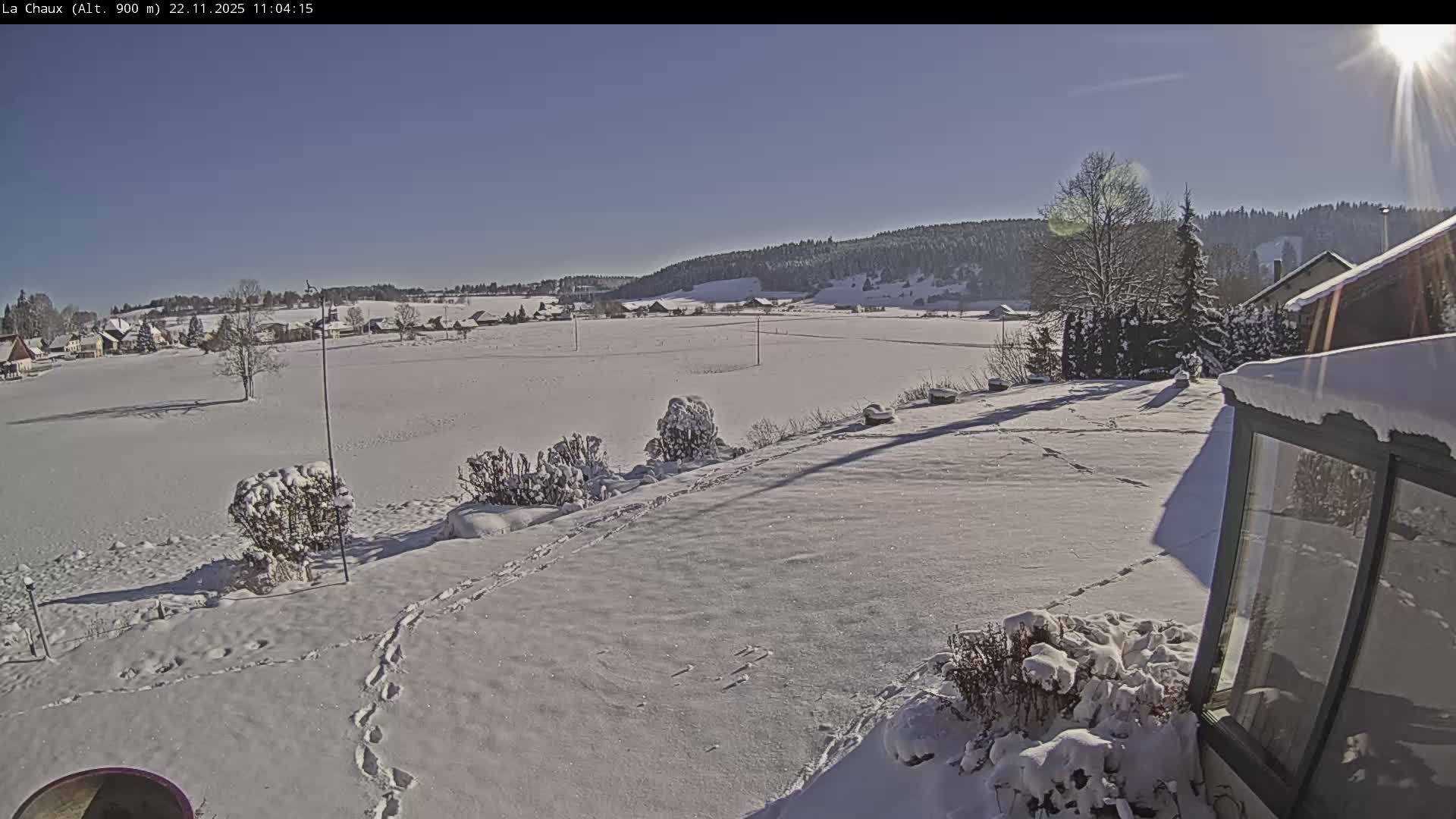 The image captures a bright, sunny winter day over a snow-covered rural landscape featuring fields, distant houses, and snow-laden bushes and structures in the foreground, all under a clear blue sky.