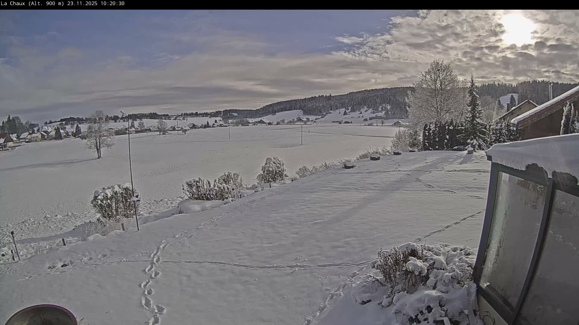 The image displays a serene, snow-covered winter landscape under a partly cloudy sky with visible sun, featuring a valley with distant scattered houses and forested hills, and a snowy foreground with bushes and footprints.