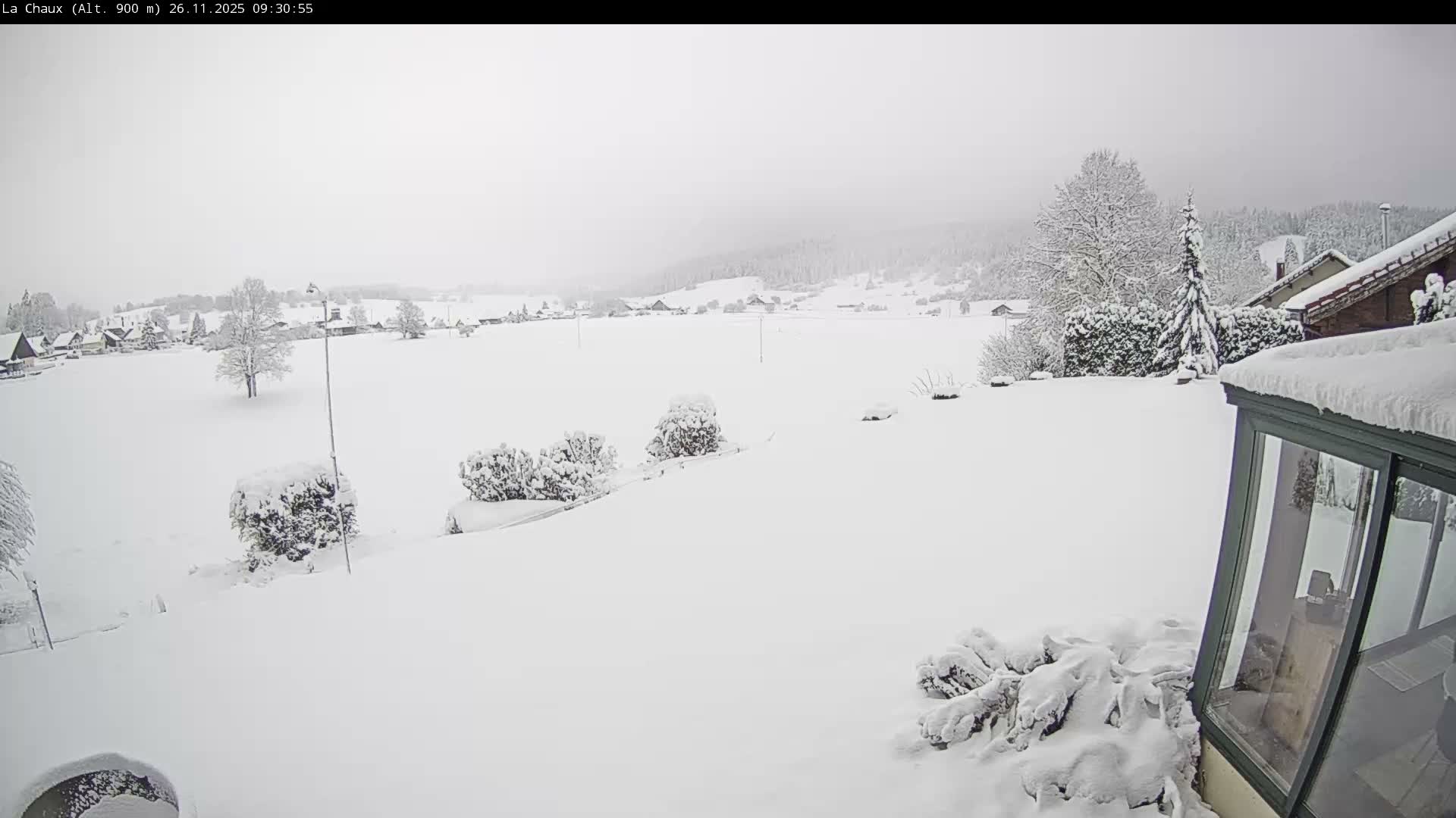 A serene rural landscape is completely blanketed in deep white snow, covering fields, trees, and distant village houses under a heavily overcast and dull winter sky.