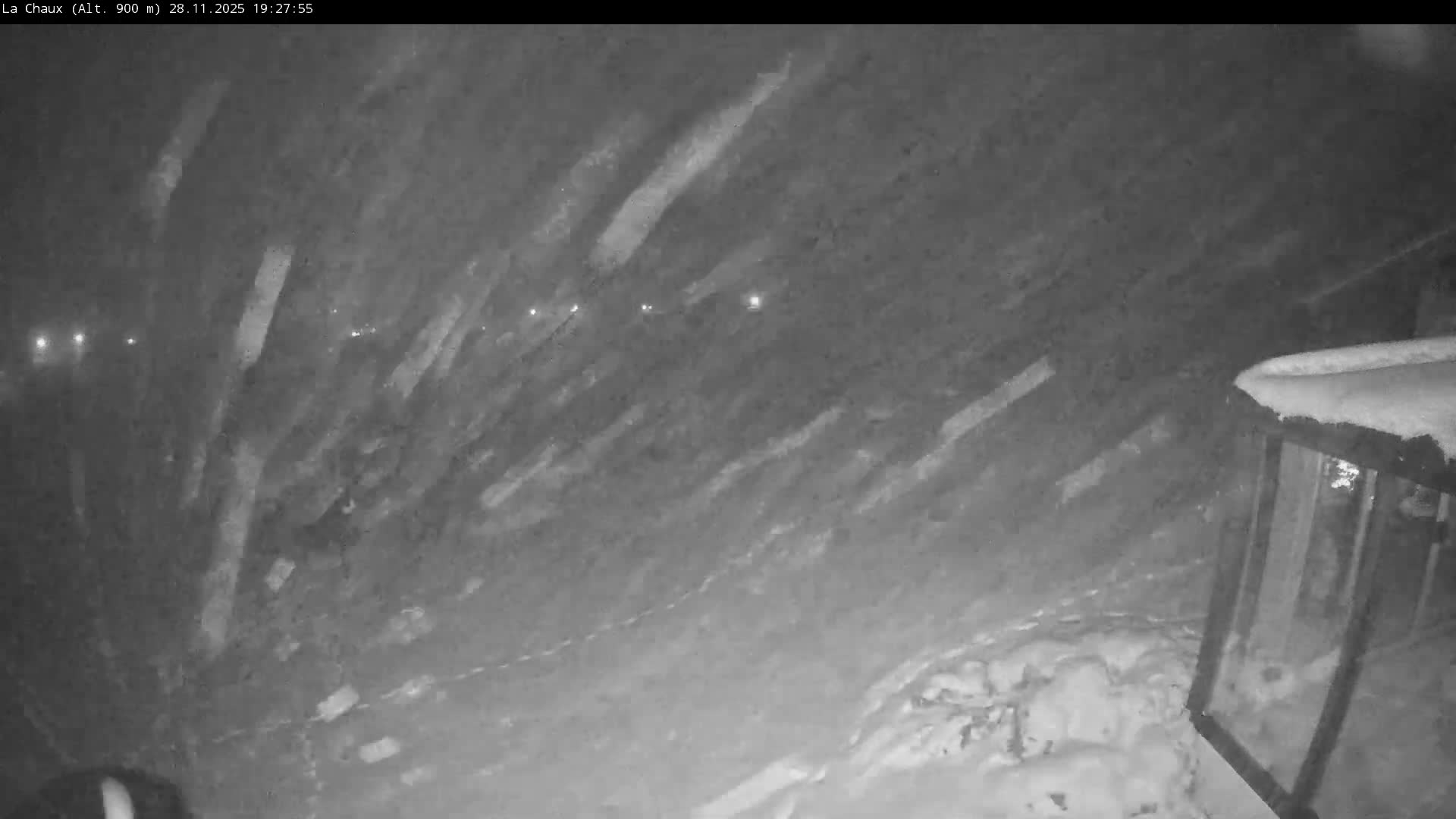 The image shows a dark, snowy outdoor scene at night with numerous blurry streaks indicating heavy snowfall, illuminated by scattered distant lights and revealing snow-covered ground and a partial structure on the right.
