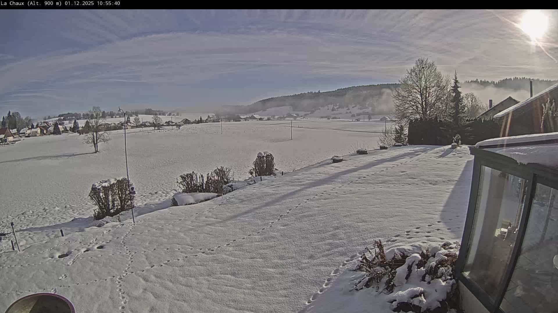 On a sunny and cold winter day, snow blankets the landscape, from footprints in the foreground to vast fields, a distant village, and forested hills with a hazy horizon, all under a partly cloudy sky.