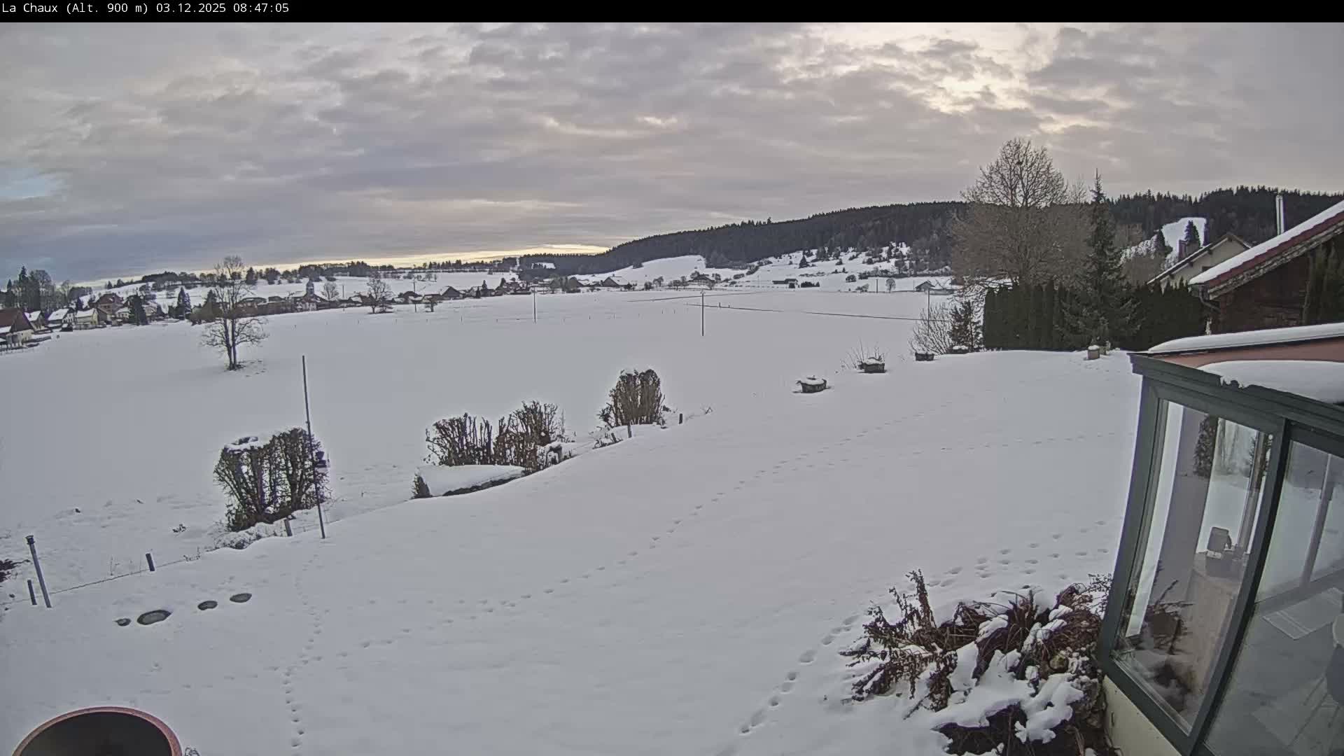 A wide view of a snow-covered winter landscape shows a village and fields extending to forested hills, with footprints marking the snowy foreground under a cloudy, overcast sky.