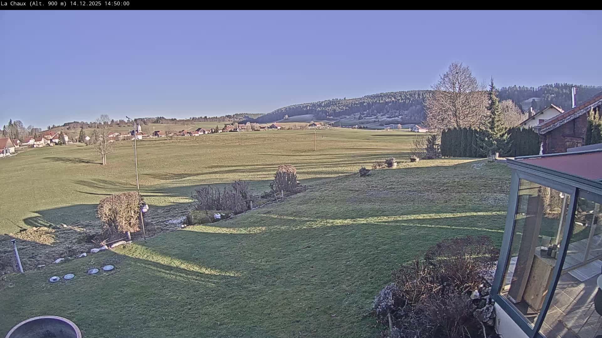 A wide view of a snow-covered winter landscape shows a village and fields extending to forested hills, with footprints marking the snowy foreground under a cloudy, overcast sky.