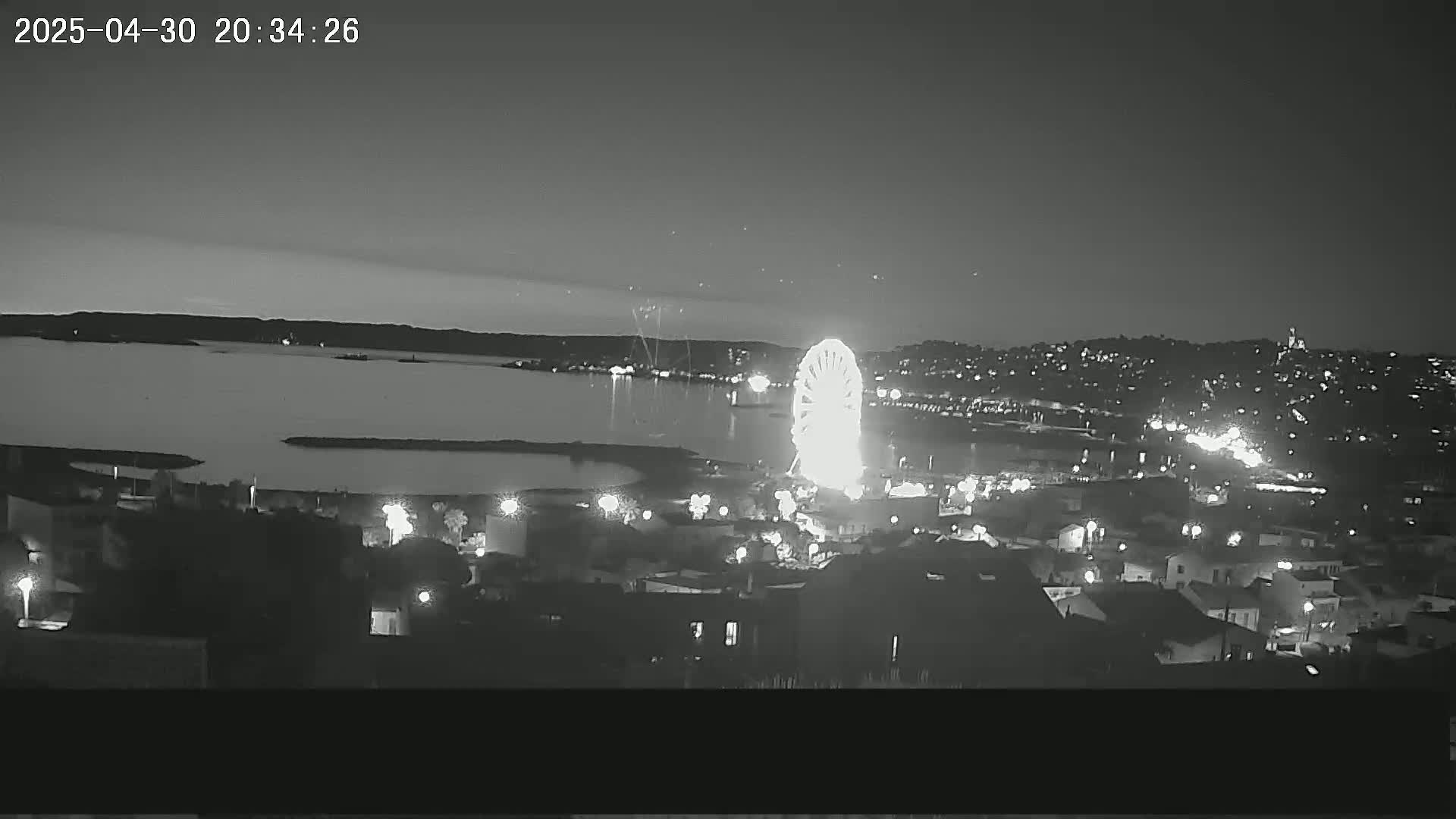 A brightly lit Ferris wheel is visible on a nighttime waterfront, overlooking a calm body of water and a city illuminated with streetlights under a dark sky.