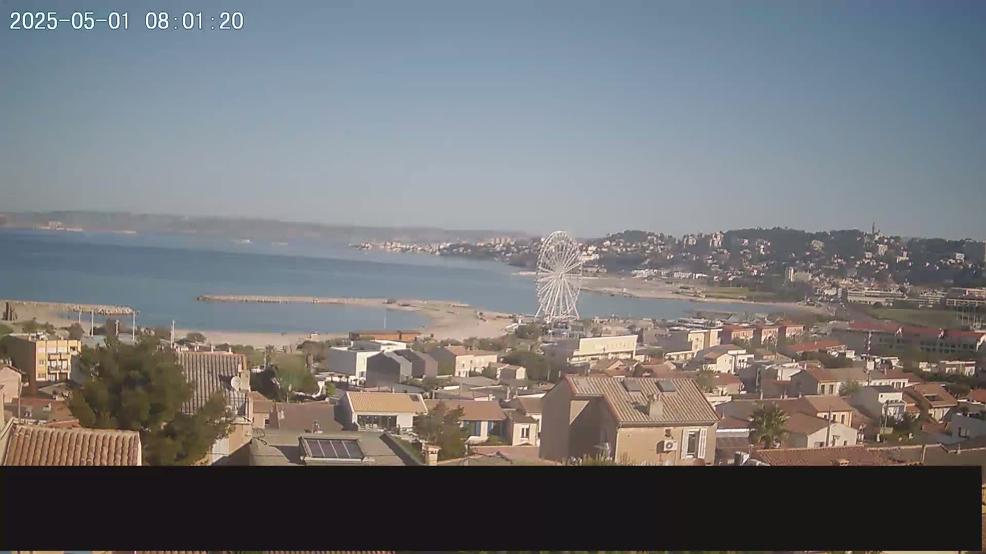 An aerial view shows a coastal town on a clear day, with a beach, calm water, and a Ferris wheel visible in the distance.