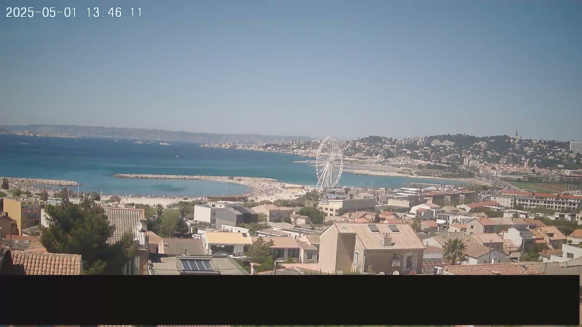 A coastal town with a Ferris wheel is seen from an elevated vantage point on a sunny day, overlooking a crowded beach and calm blue sea.