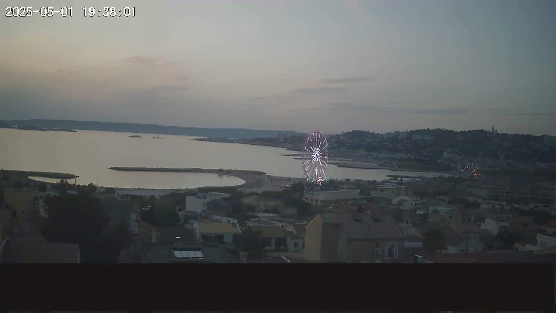 A brightly lit Ferris wheel stands near a calm bay at dusk, overlooking a coastal town.
