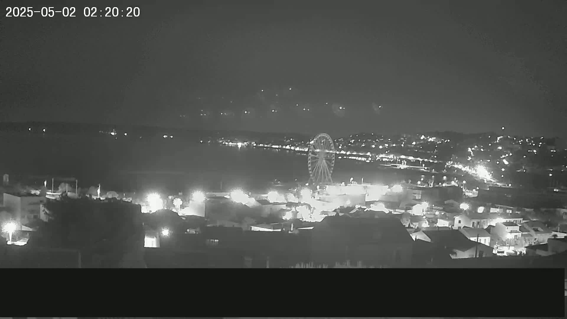A nighttime, high-angle view of a coastal town with a Ferris wheel near a body of water, under a clear sky.