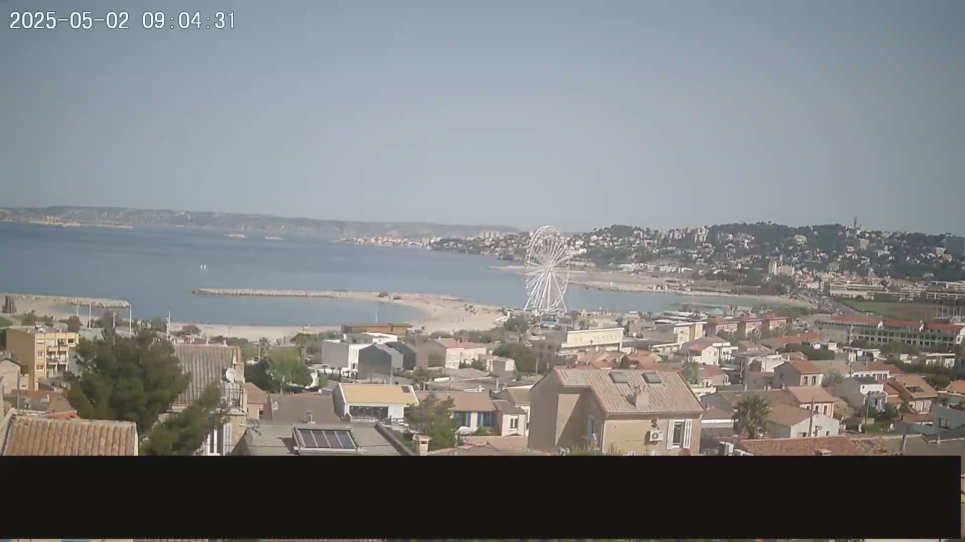 A coastal town with a Ferris wheel is visible on a sunny day, overlooking a calm bay and distant hills.