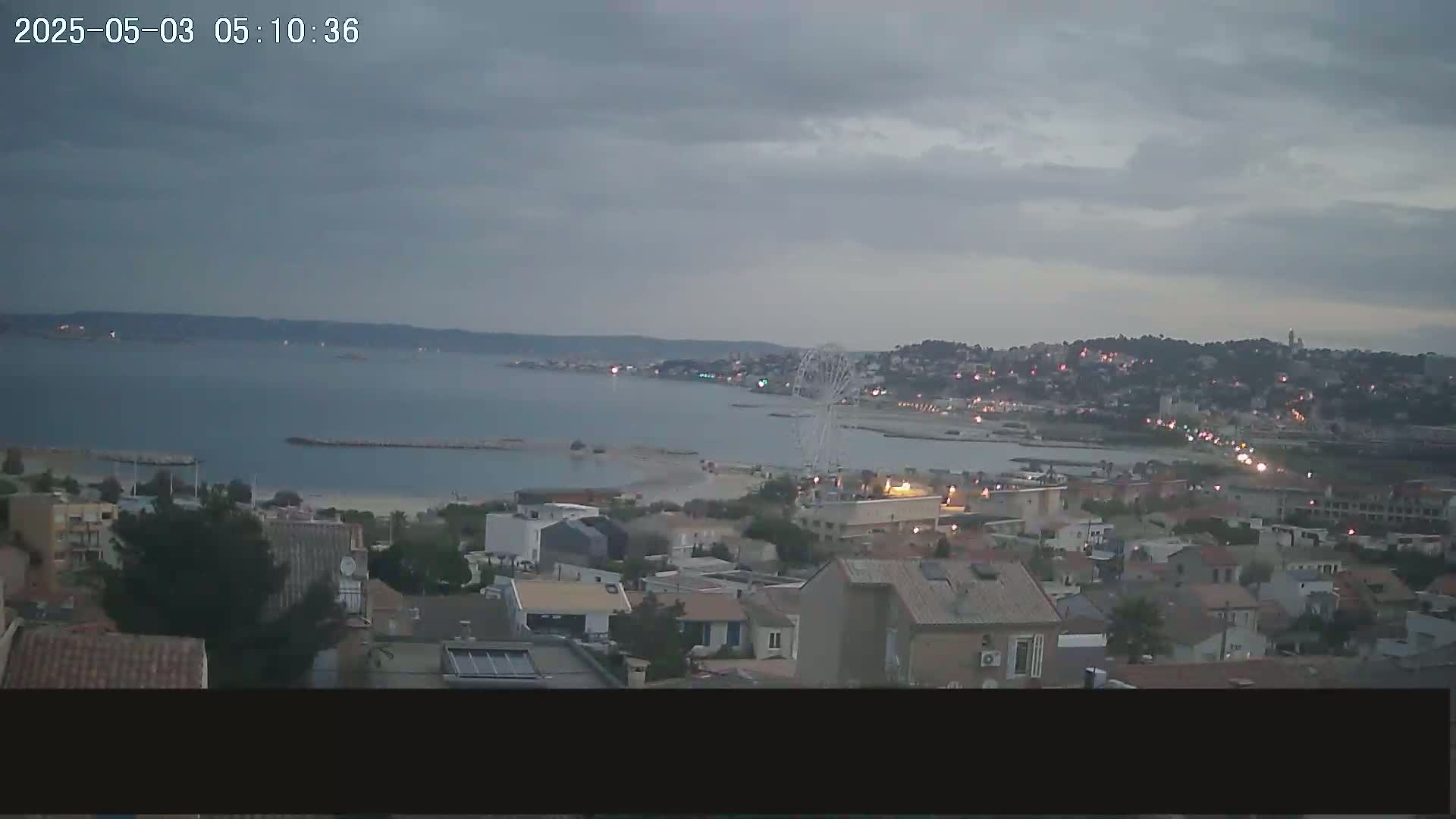 An overcast sky overlooks a coastal town with a Ferris wheel visible near the beach and calm waters.
