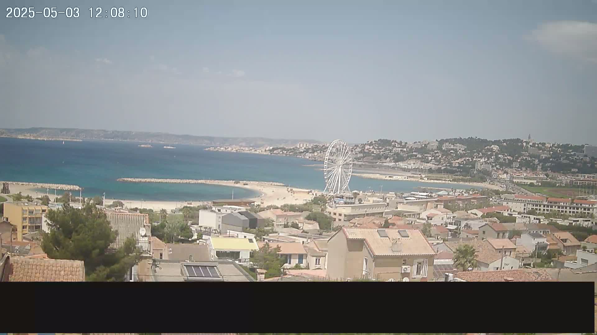 A coastal town with a Ferris wheel on a partly sunny day, overlooking a calm bay.