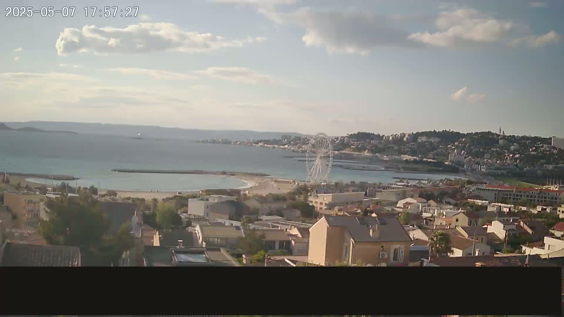 A coastal town with a Ferris wheel is seen on a partly cloudy day, overlooking a calm bay and distant hills.