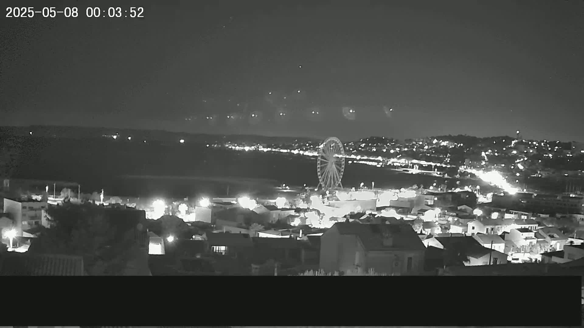 A nighttime, high-angle view of a coastal town with a Ferris wheel visible in the distance, under a clear sky.