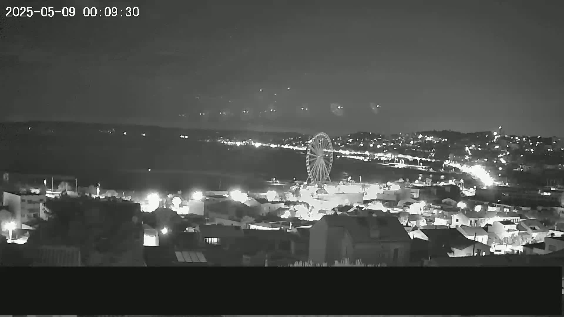 A nighttime, low-light view shows a Ferris wheel and a city illuminated with lights under a clear sky.