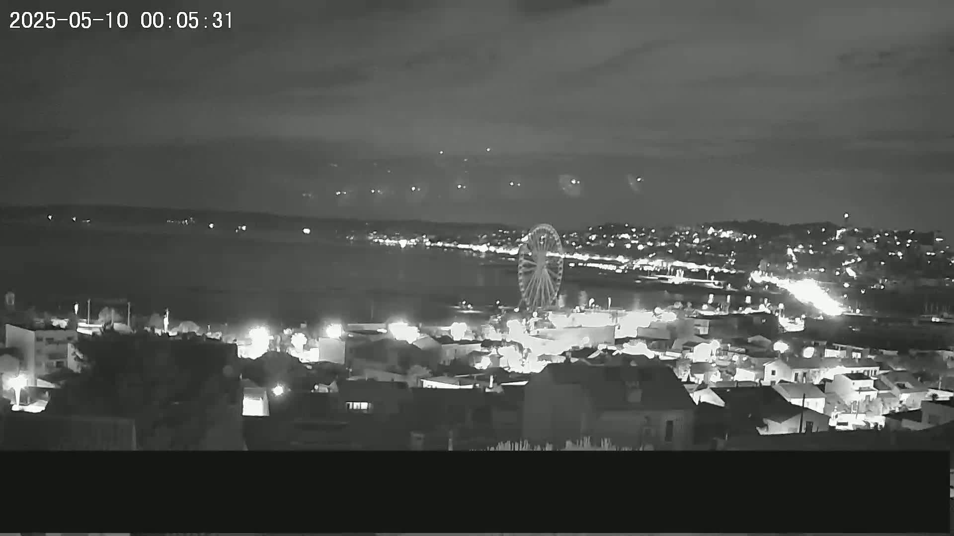 A nighttime, high-angle view shows a Ferris wheel and a coastal town illuminated by lights, with a dark body of water in the background under a mostly clear sky.
