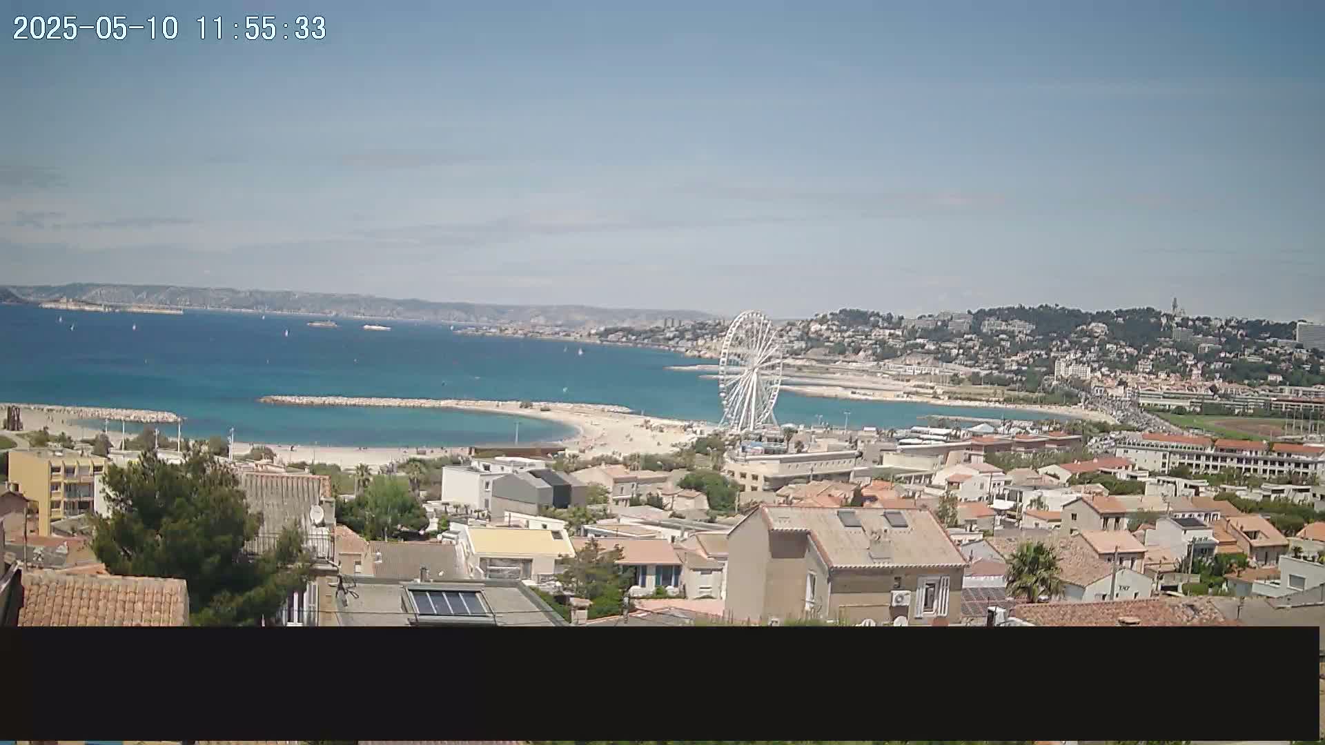 A coastal town with a Ferris wheel is visible on a sunny day, overlooking a calm bay with several sailboats.