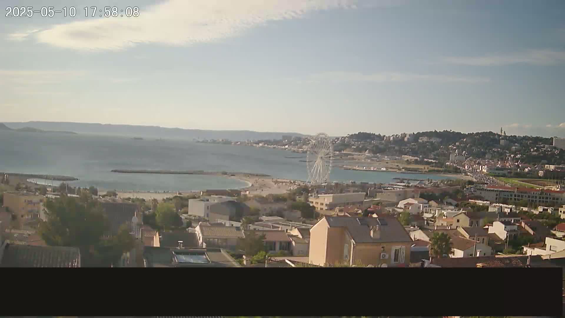 An elevated view of a coastal town on a partly sunny day shows a Ferris wheel near a beach, with calm water and distant hills visible.