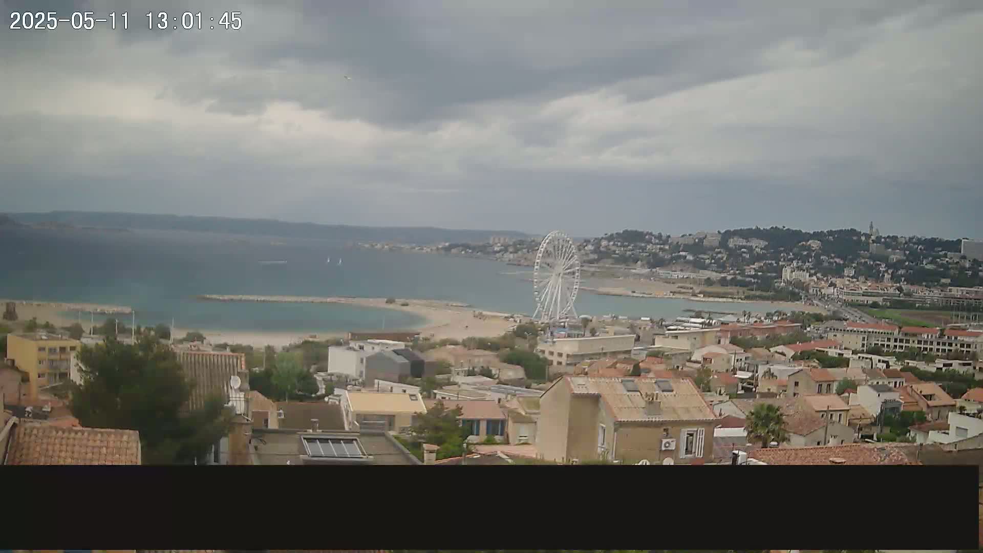 An overcast day reveals a coastal town with a Ferris wheel near a beach, viewed from a hilltop.