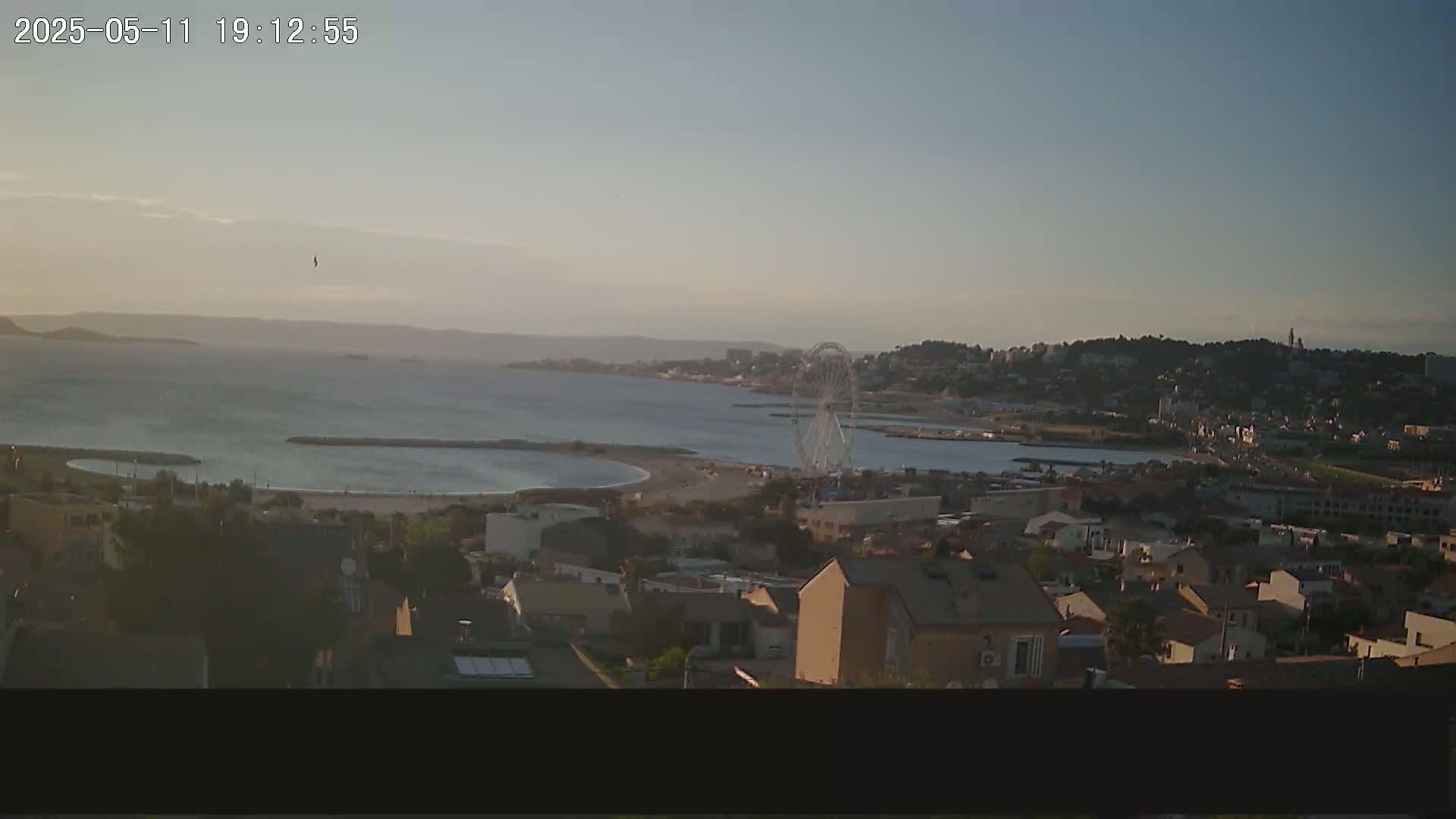 A coastal town at sunset, featuring a Ferris wheel near a calm bay, is seen from a hilltop vantage point.