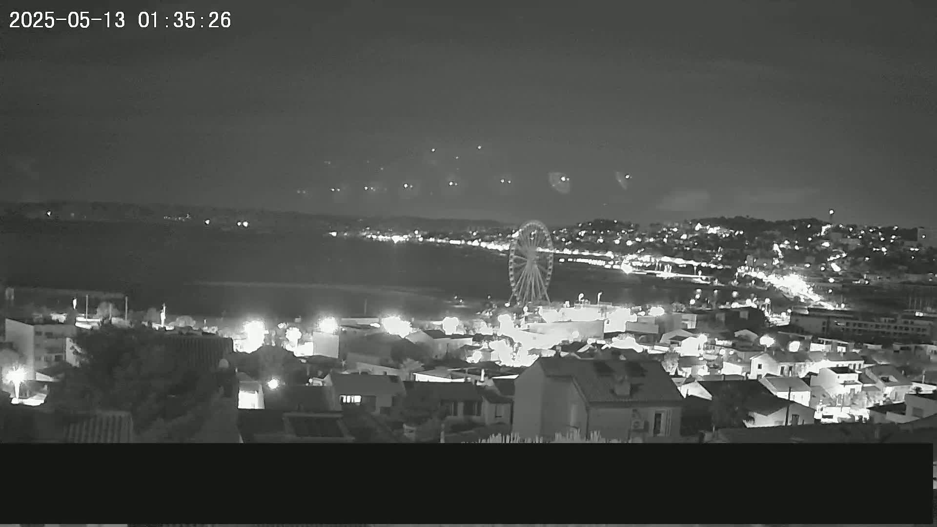 A nighttime, high-angle view shows a Ferris wheel and a town illuminated with lights along a body of water under a clear sky.