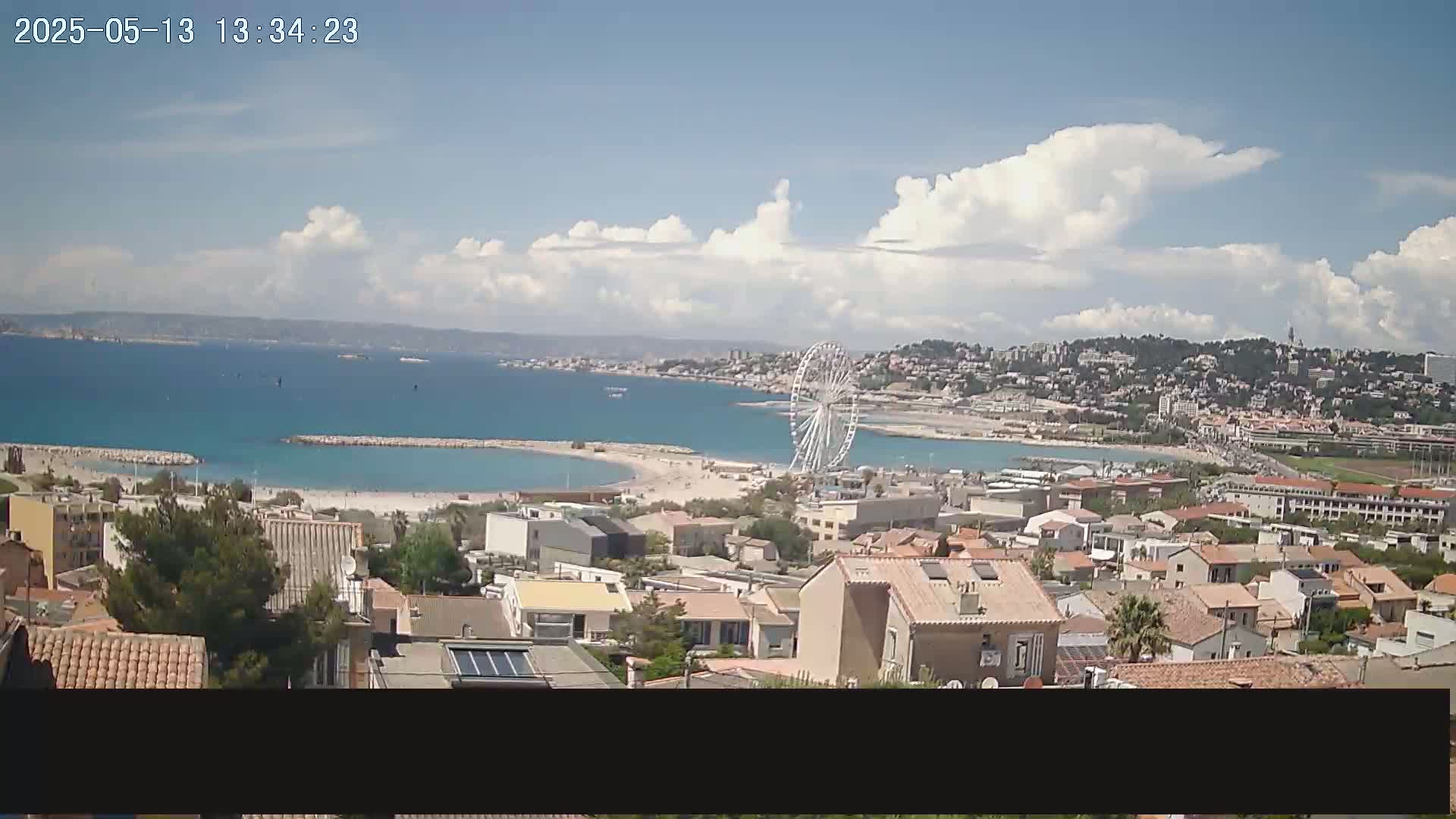 A coastal town with a Ferris wheel is visible on a partly cloudy day, overlooking a calm, turquoise bay.