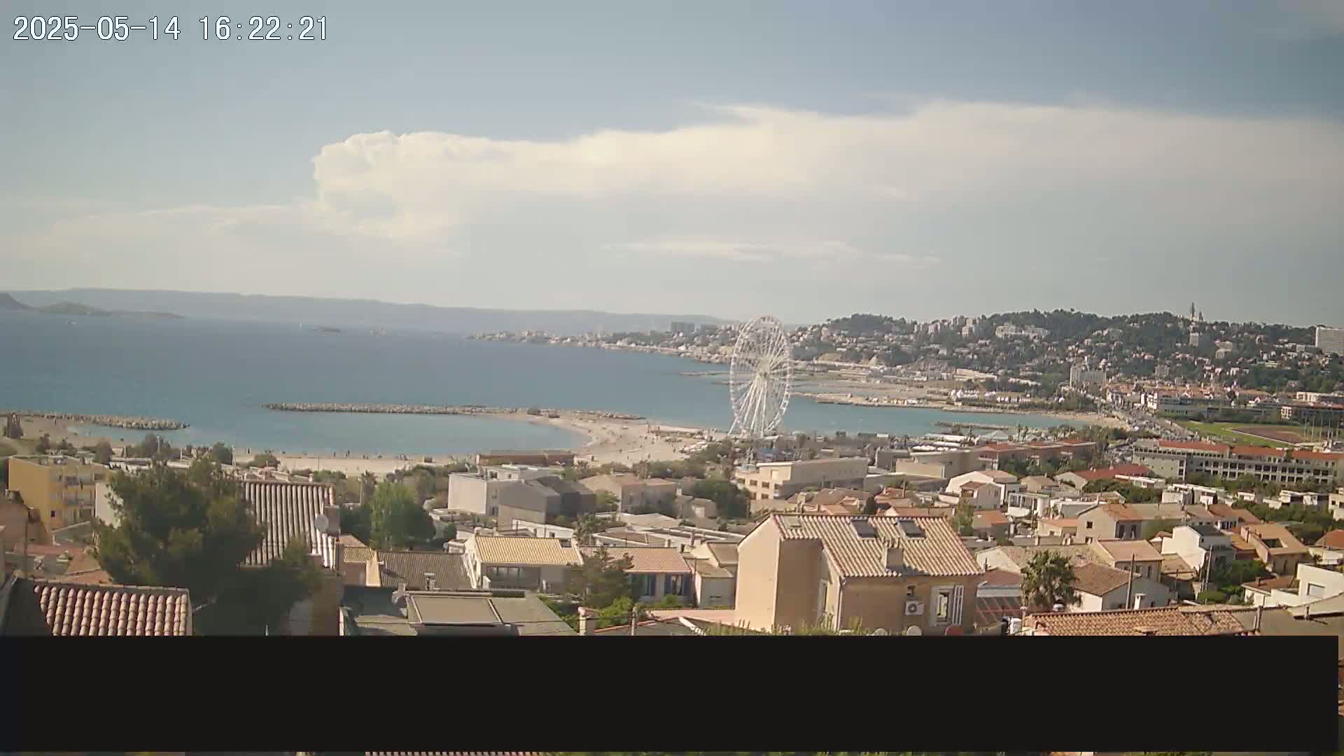 A coastal town with a Ferris wheel is visible on a partly cloudy day, overlooking a calm bay and distant hills.