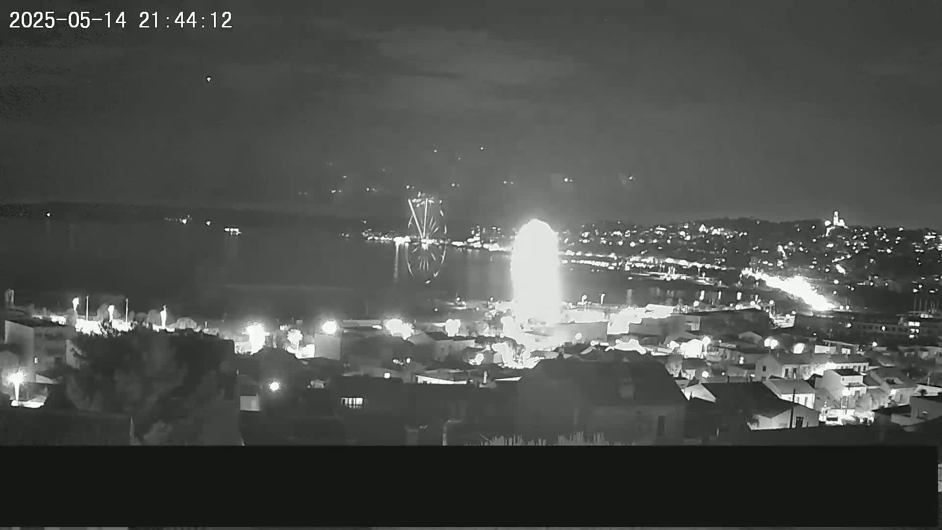 A nighttime, high-angle view shows a city by a body of water with fireworks displaying over the water and city.