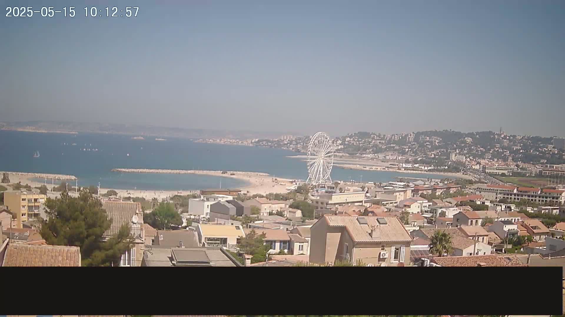 A coastal town on a sunny day with a Ferris wheel visible near a sandy beach and calm sea.