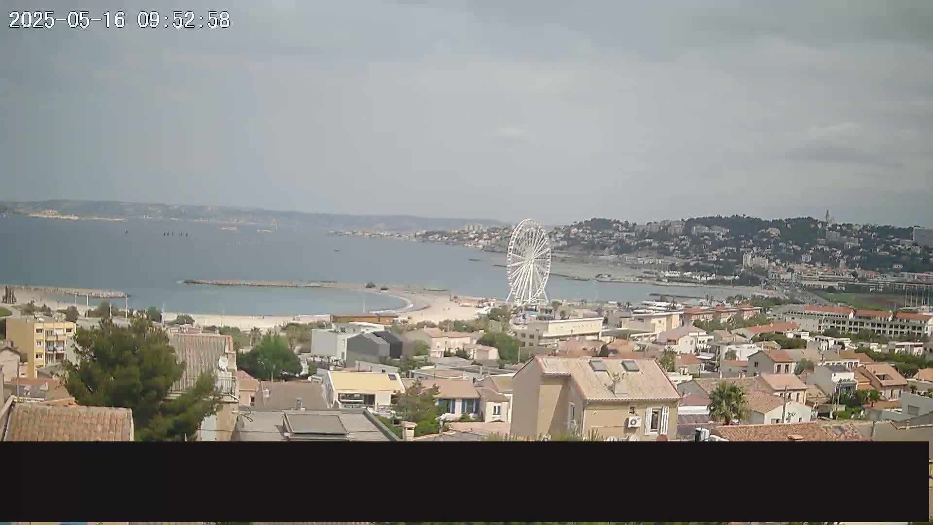 A coastal town with a Ferris wheel is seen on a partly cloudy day, overlooking a calm bay.