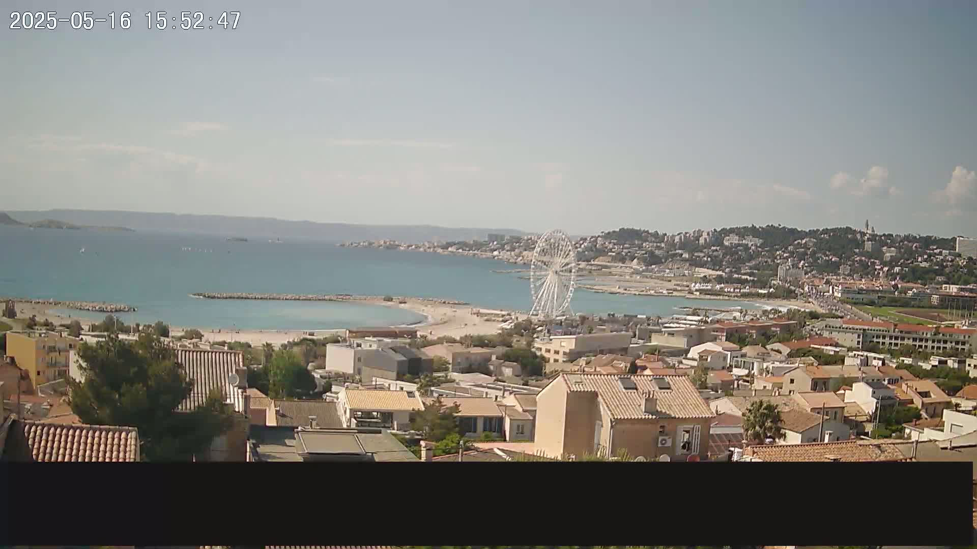 A coastal town on a sunny day with a Ferris wheel near a sandy beach and calm ocean waters, viewed from a hilltop overlooking a collection of buildings.