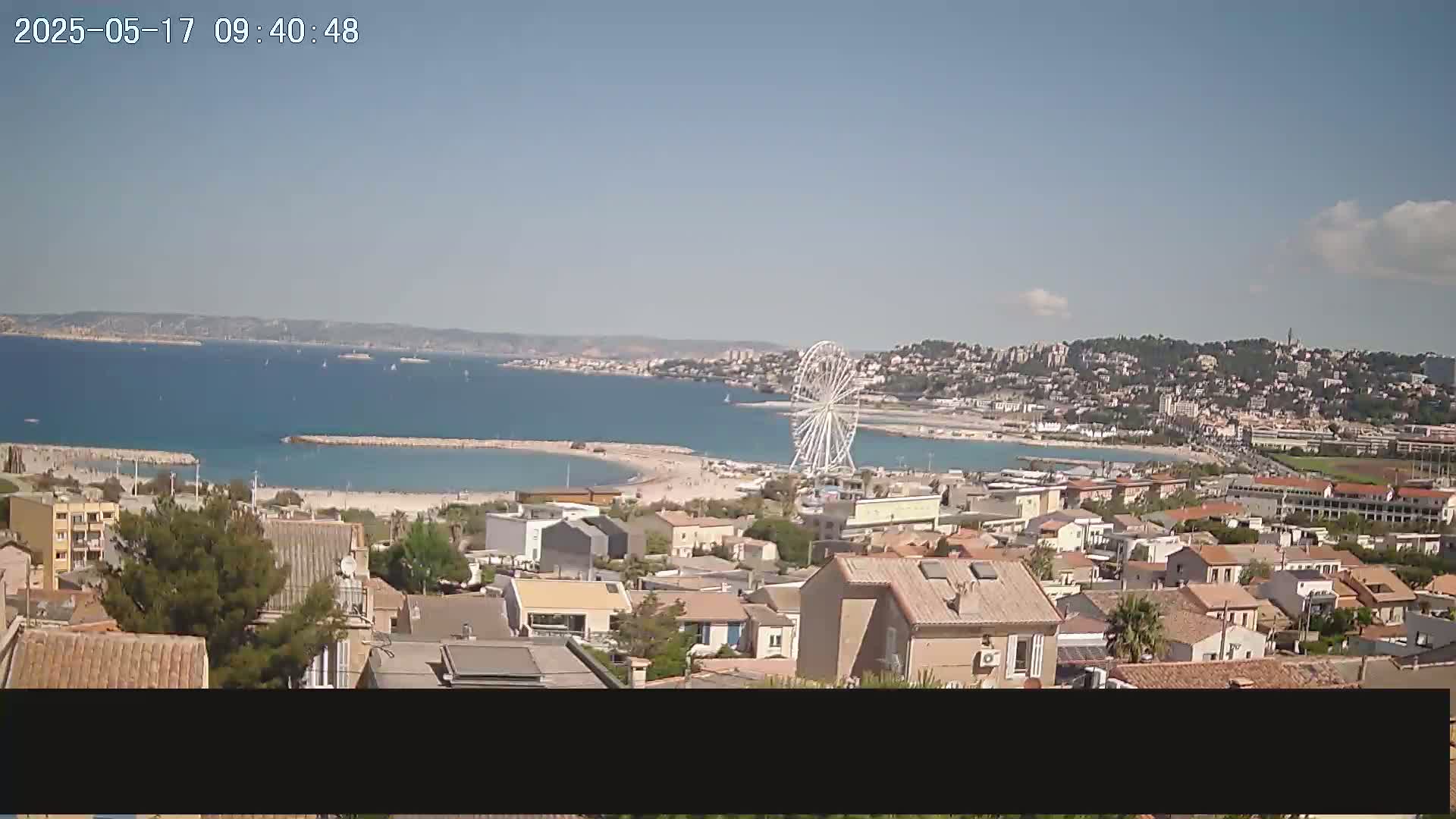 A coastal town with a Ferris wheel is visible, overlooking a calm bay with a sandy beach on a sunny day.