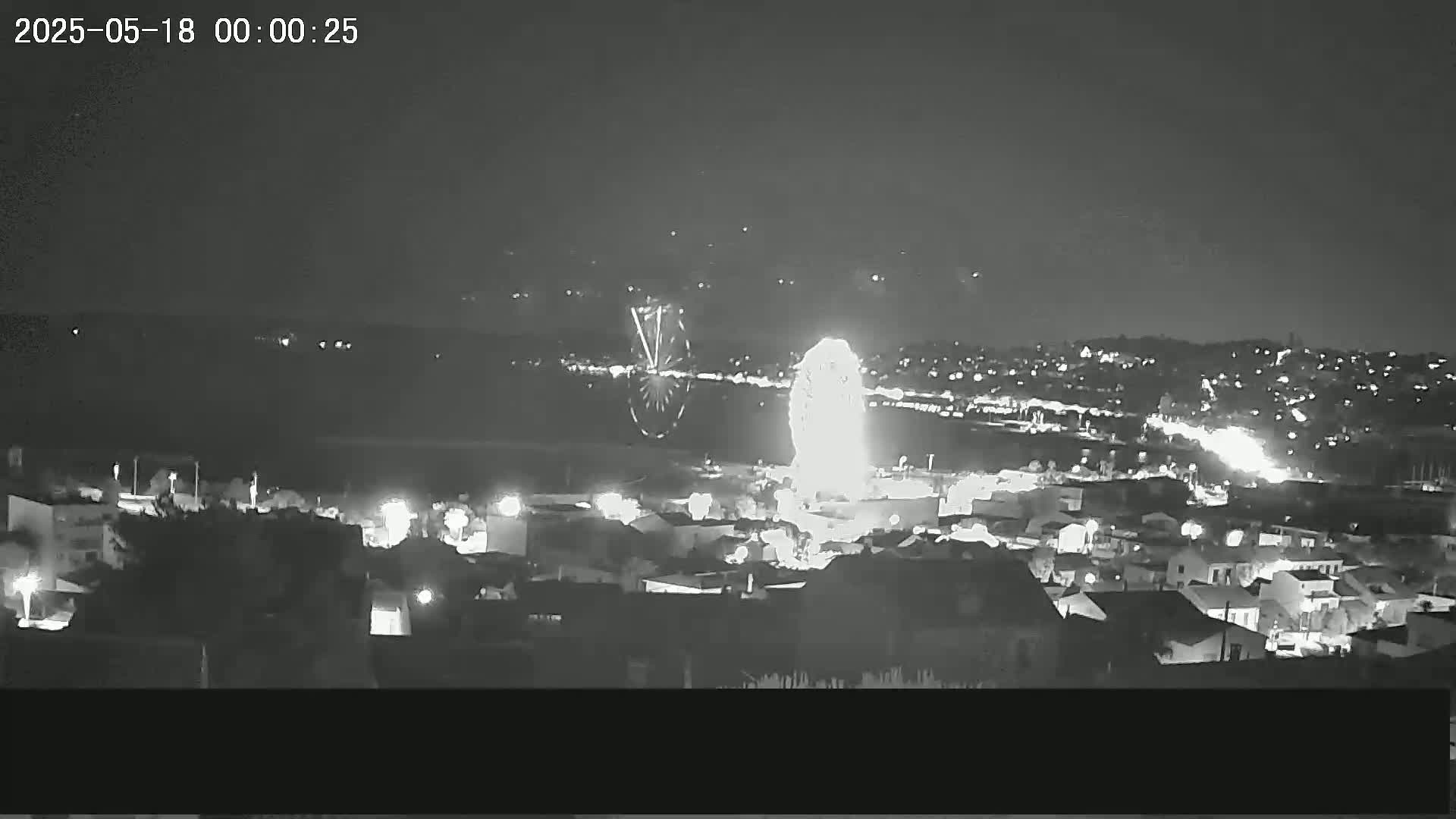 A nighttime, long-range view of a town with fireworks exploding over a Ferris wheel.