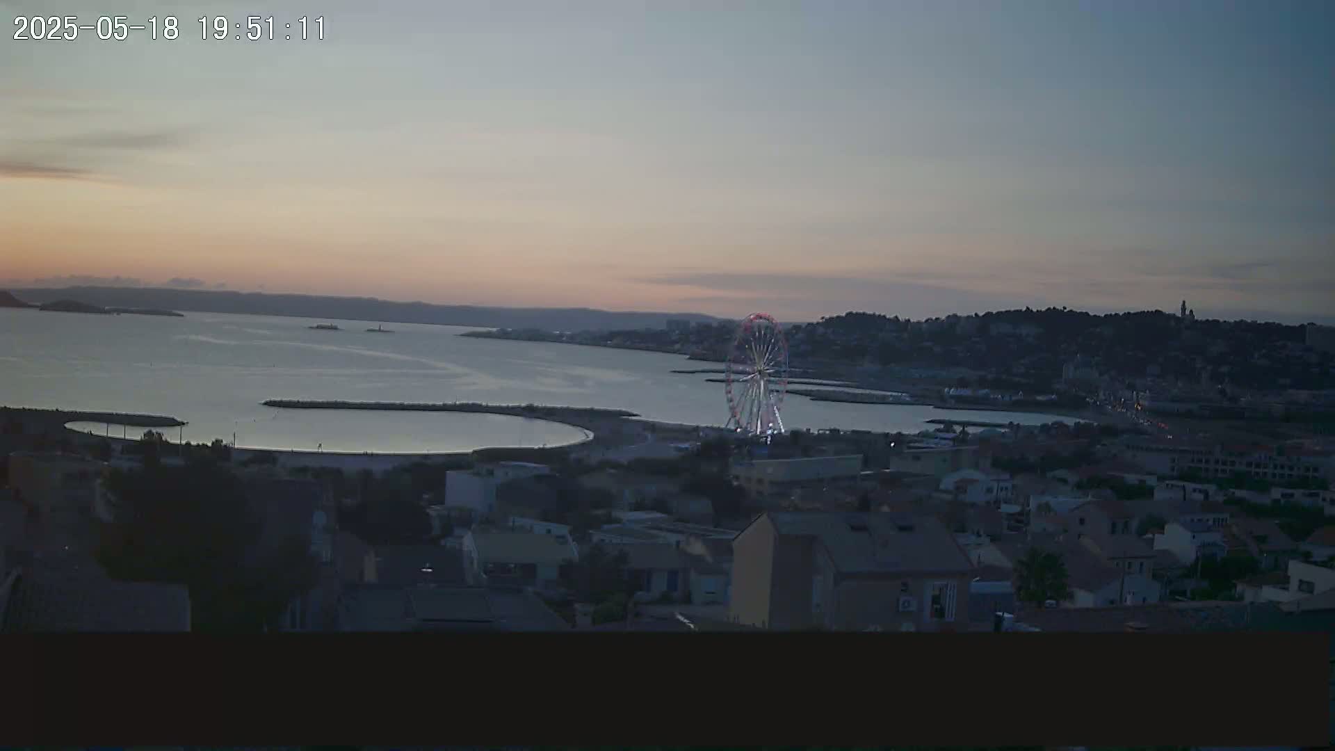 A coastal town at dusk, with a Ferris wheel near the water, under a mostly clear sky.