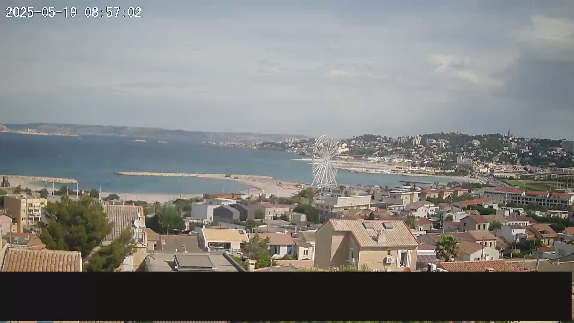 A coastal town with a Ferris wheel is visible on a partly cloudy day, overlooking a calm bay.