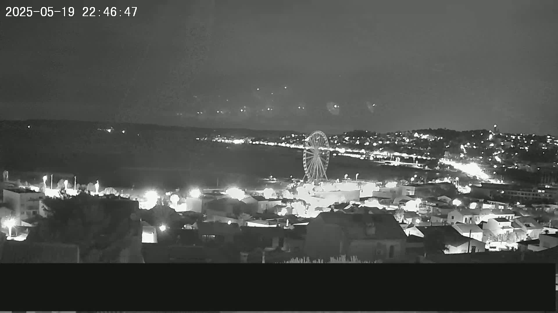 A nighttime view of a coastal town with a Ferris wheel on the waterfront, under a clear sky with faint lights possibly from fireworks in the distance.