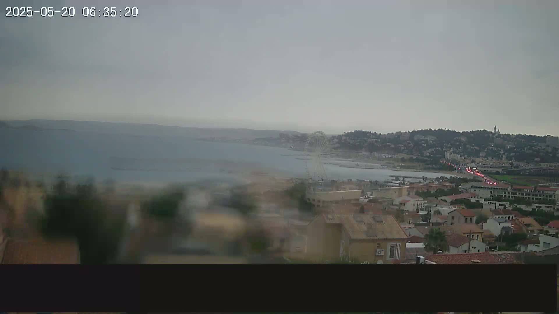 A rainy, overcast view of a coastal town with a Ferris wheel visible across the bay, showing buildings and a road with traffic.