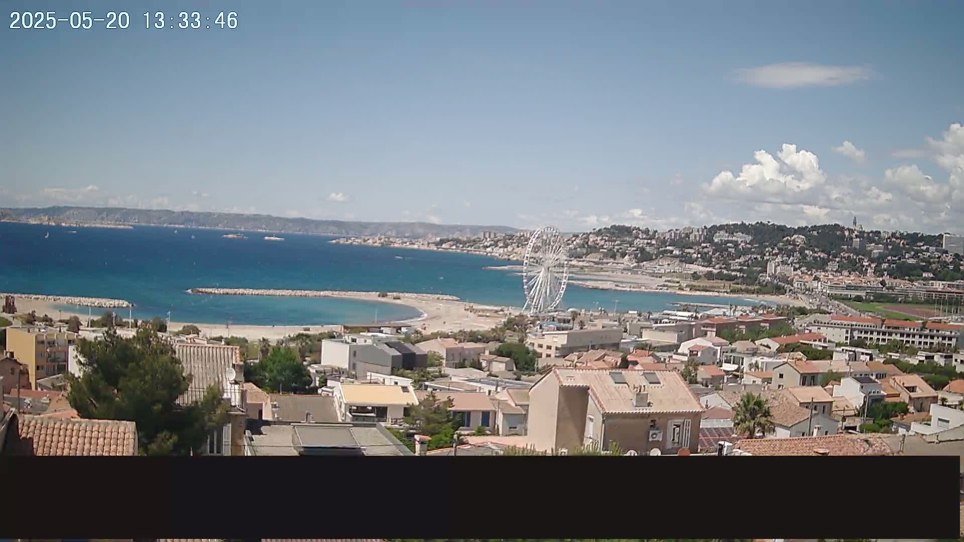 A coastal town with a Ferris wheel is visible on a partly cloudy day, overlooking a calm bay and distant mountains.