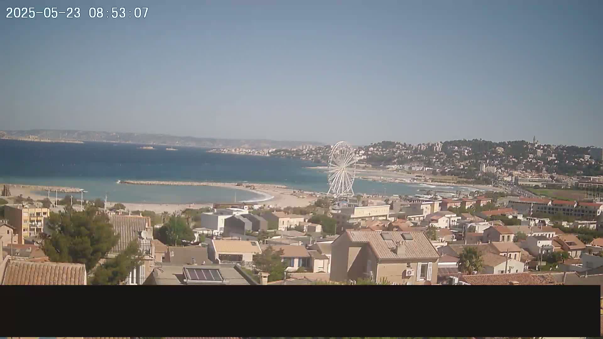 A coastal town with a Ferris wheel overlooks a calm, light-blue sea under a clear sky.