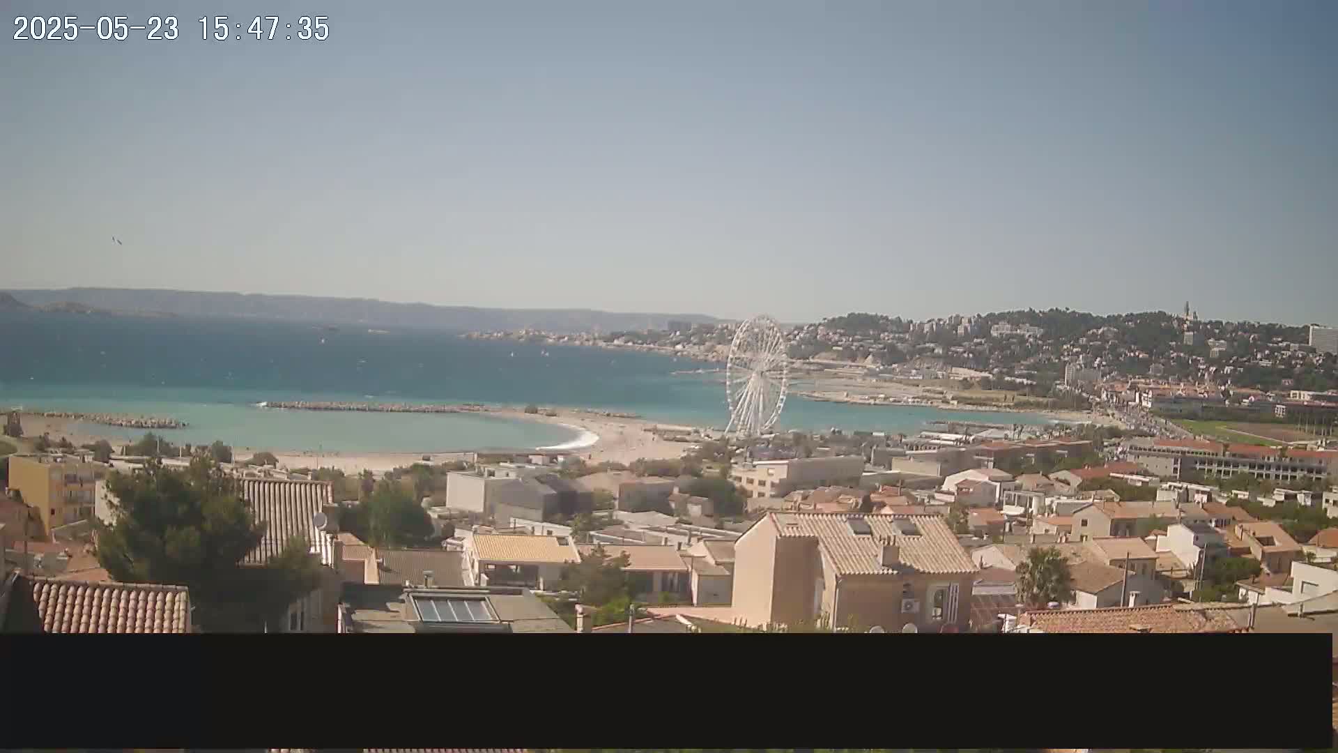A coastal town with a Ferris wheel is visible on a sunny day, overlooking a calm, turquoise bay.