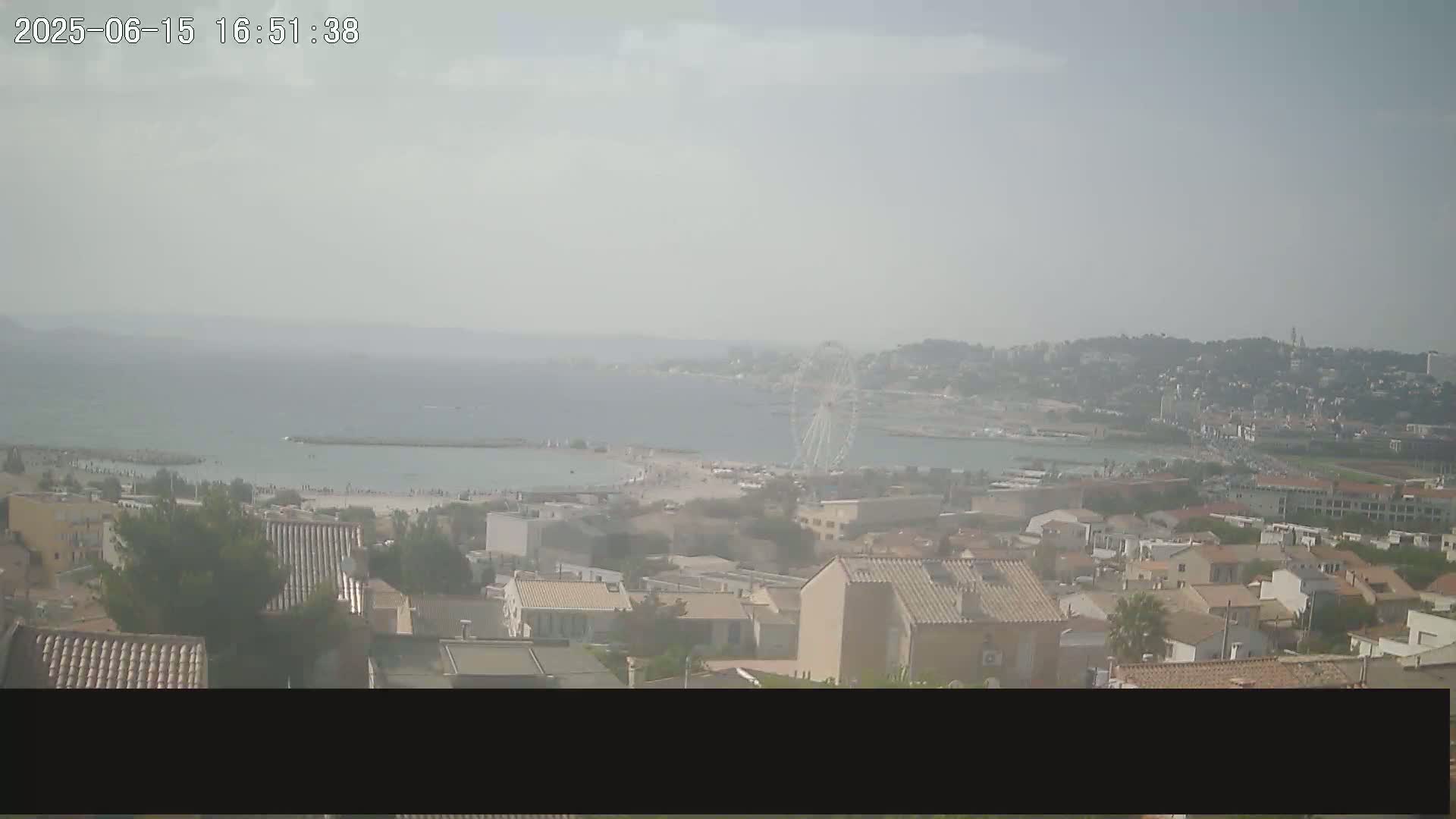 A hazy view shows a coastal town with a beach and calm sea, a Ferris wheel in the distance.