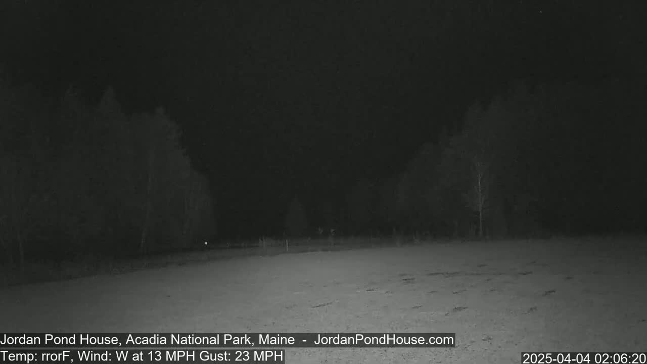 A nighttime, low-light view of a flat, snow-covered area with sparsely spaced trees in the background under a dark sky, with a westerly wind at 13 mph and gusts up to 23 mph.