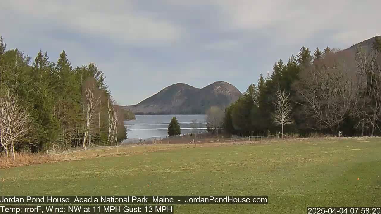 A grassy field overlooks a partly frozen lake and two rounded mountains under a cloudy sky.