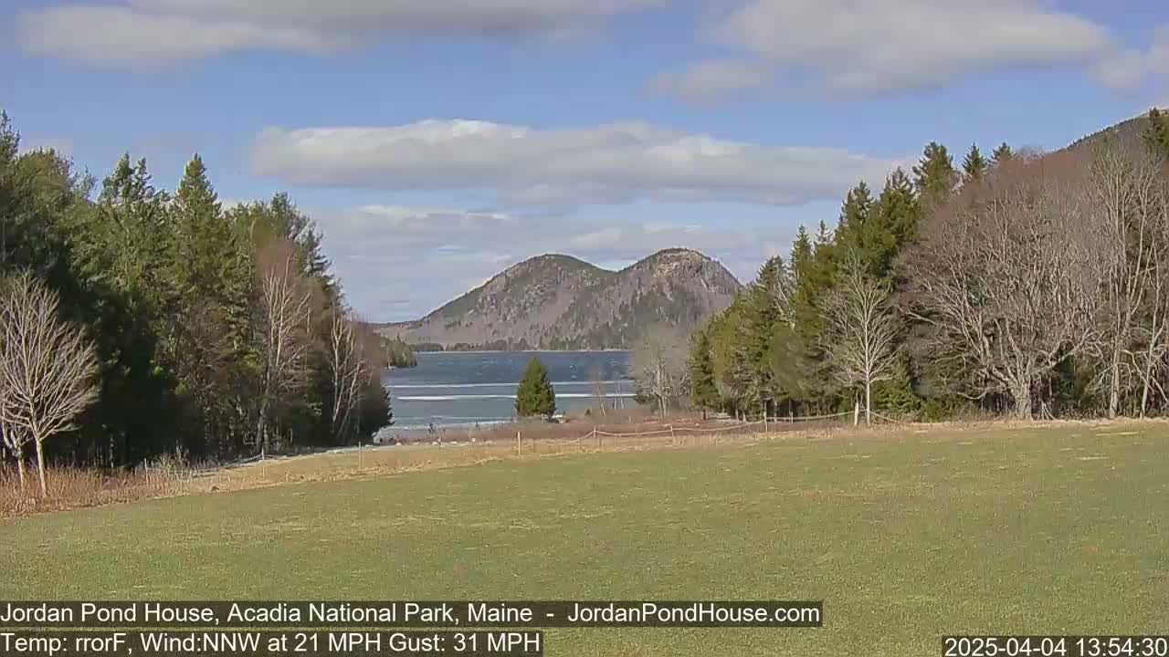 A partly sunny day shows a calm lake nestled between two rocky mountains, framed by leafless and budding trees, and a grassy field in the foreground.