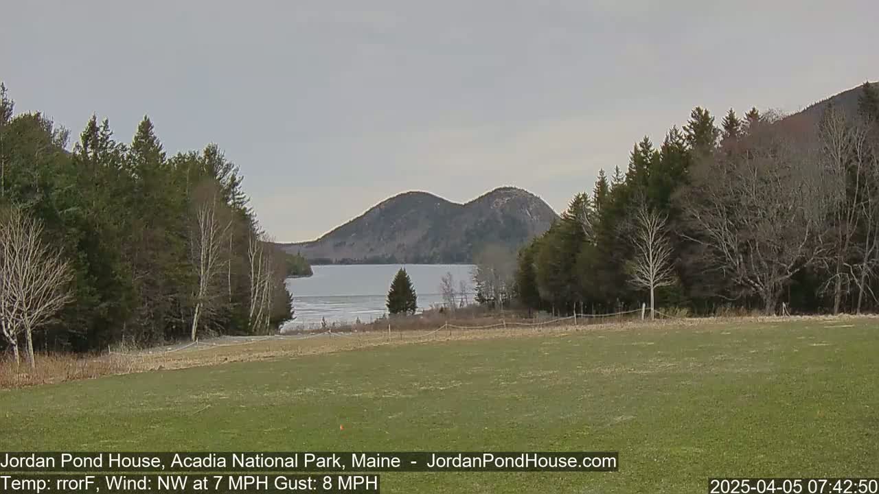 A partly frozen lake is nestled between two rounded mountains under a cloudy sky, viewed from a grassy field bordered by bare and evergreen trees.