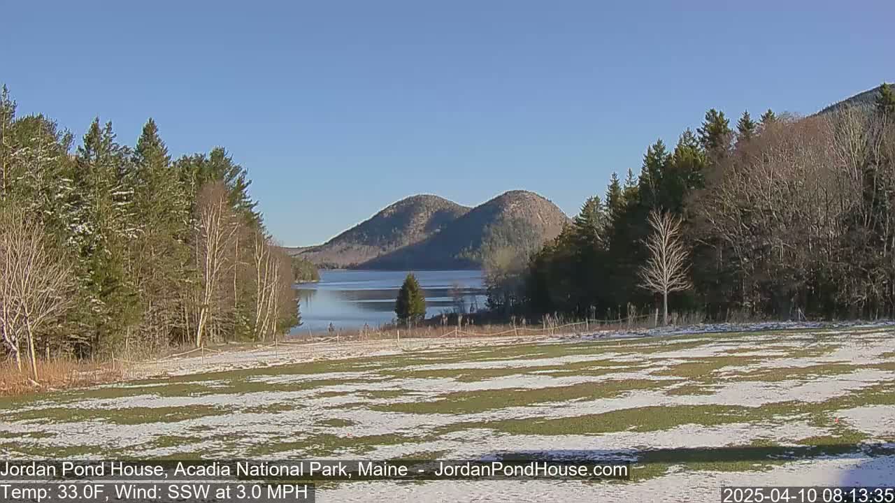A snow-dusted field overlooks a calm lake nestled between two rounded mountains under a clear blue sky.