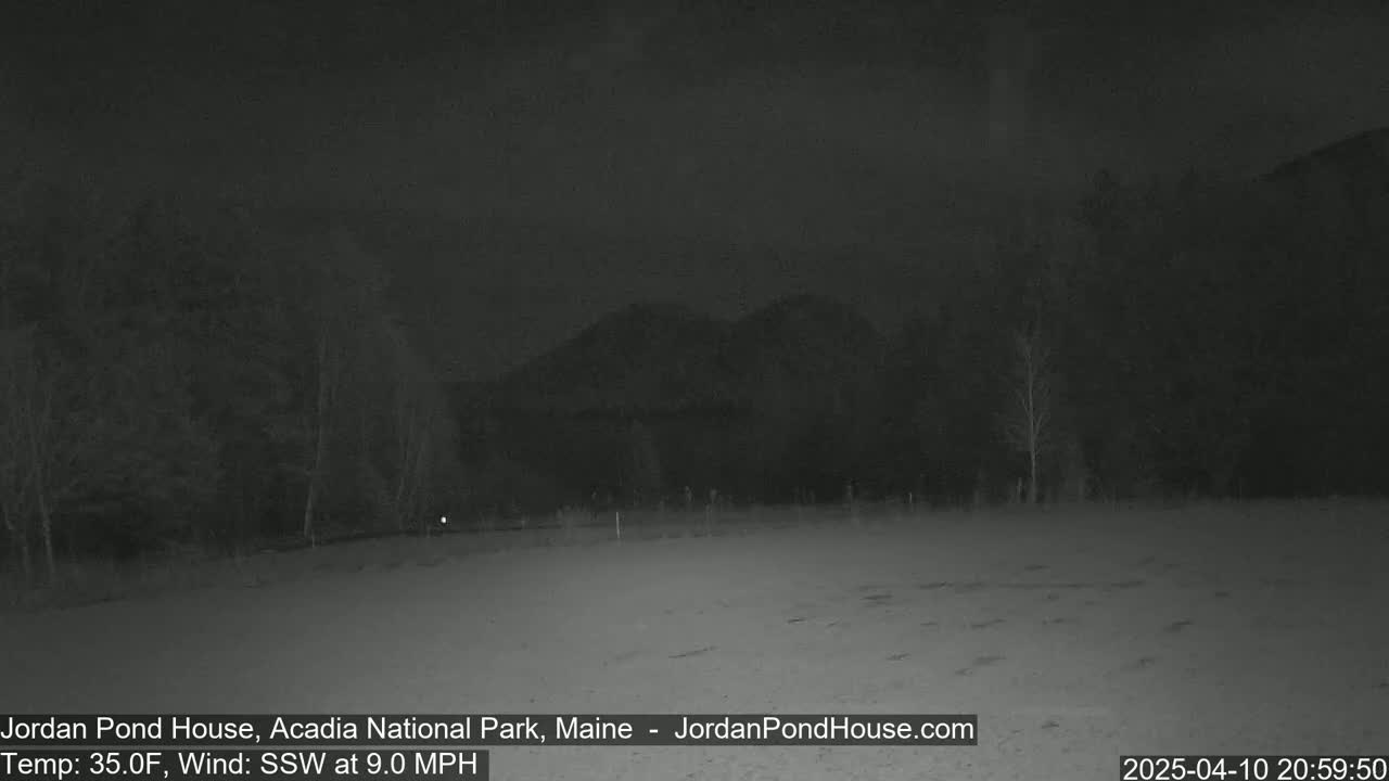 A dark, nighttime scene shows a flat, snow-covered field with trees and a hill in the background under a mostly clear sky, with a 9 mph wind from the south-southwest.