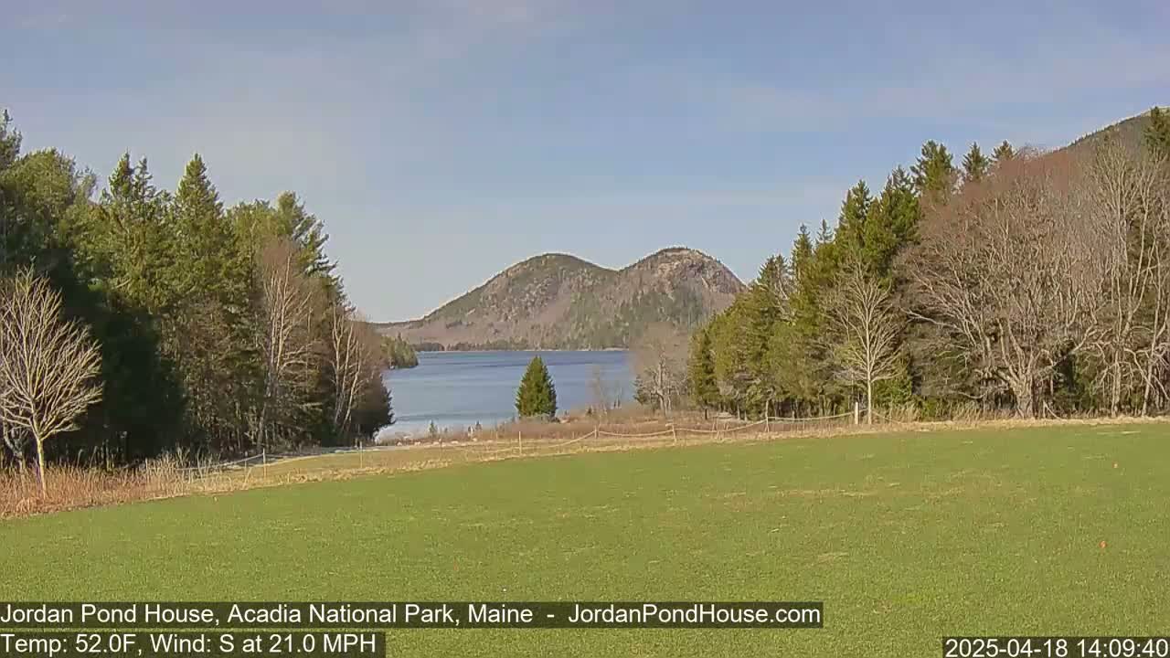 A calm, sunny day reveals a tranquil lake nestled between verdant hills and surrounded by early spring trees, with a grassy field in the foreground.