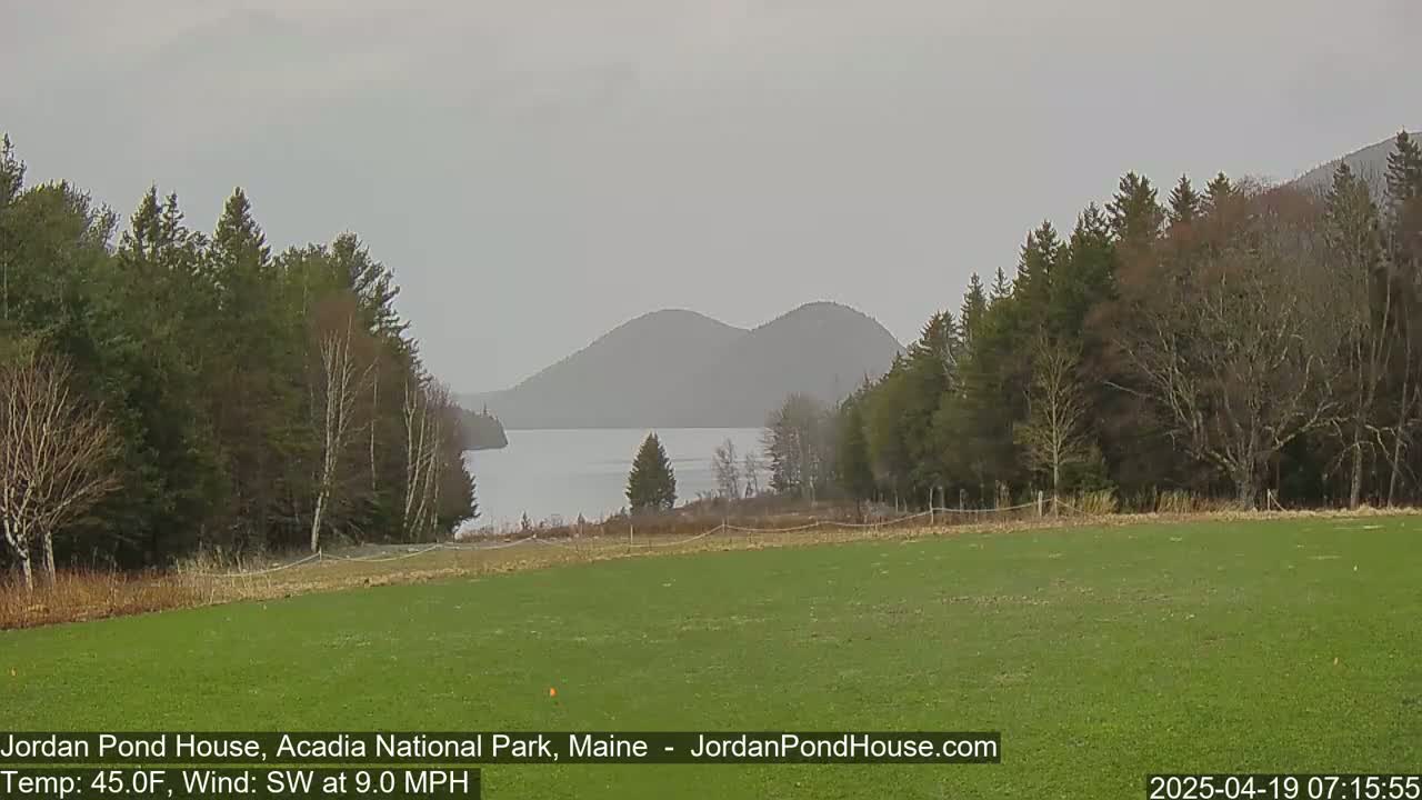 A grassy field overlooks a calm lake and distant mountains under an overcast sky.