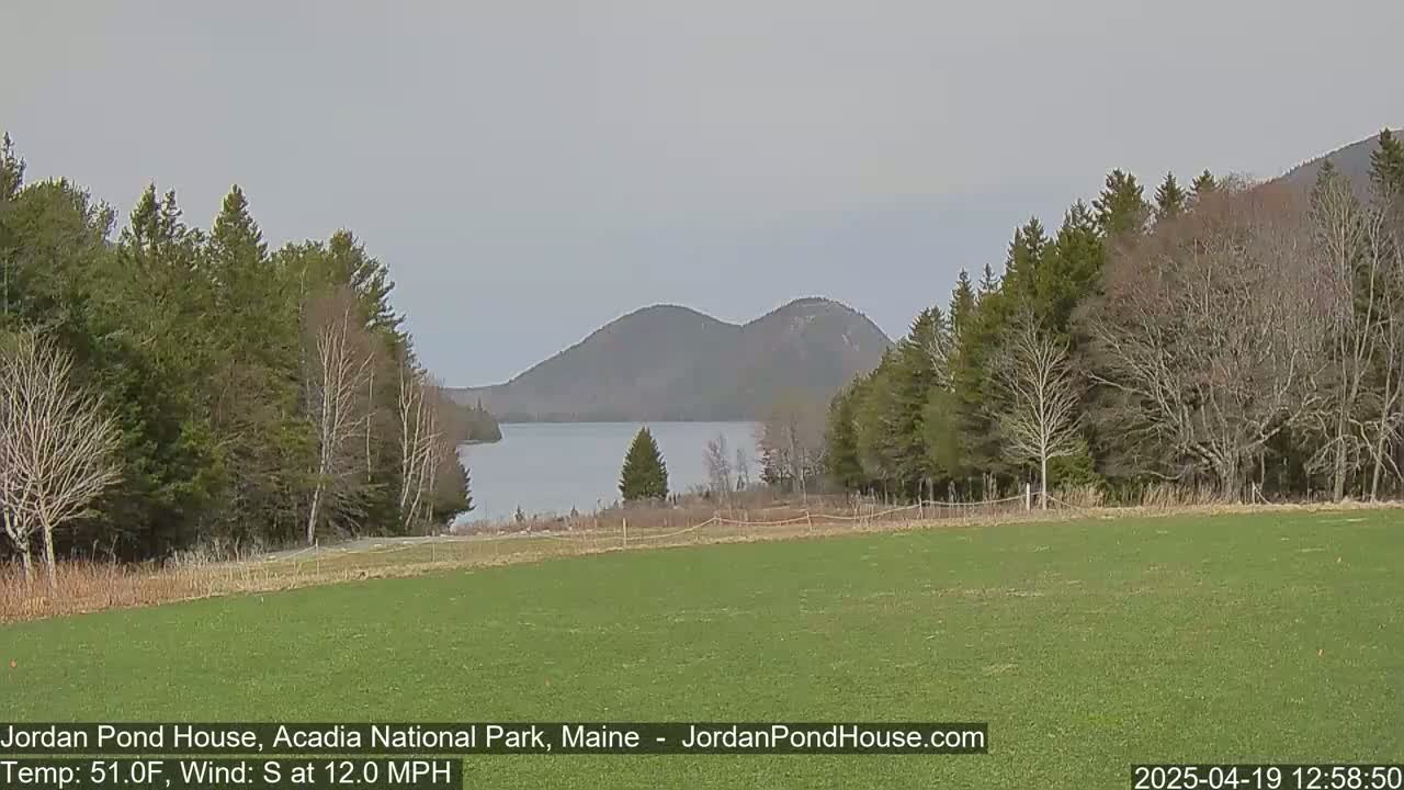 A grassy field overlooks a calm lake nestled between wooded hills under a mostly cloudy sky, with a southerly wind blowing at 12 mph.
