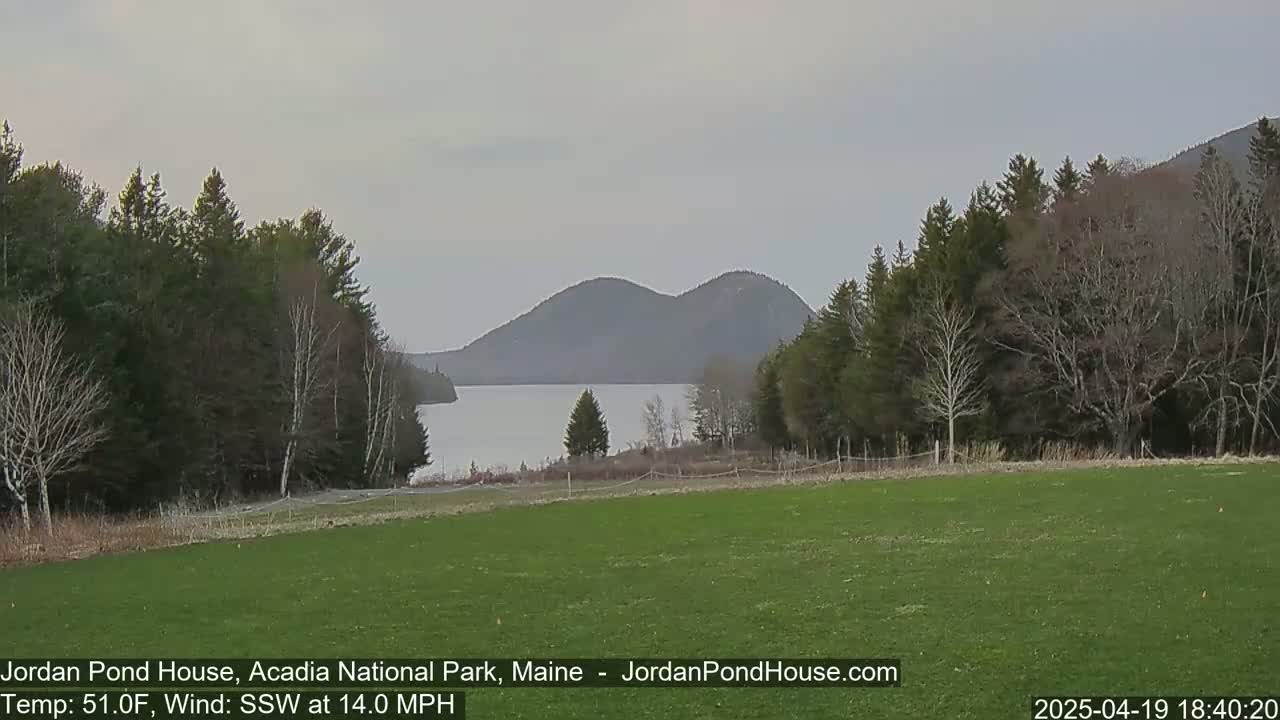 A grassy field overlooks a calm lake and distant mountains under an overcast sky, with a moderate south-southwesterly wind.